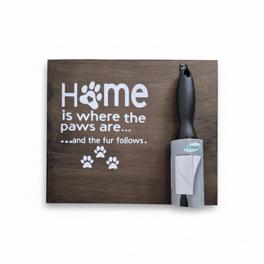 Rustic wooden decorative sign with white text reading "Home is where the paws are... and the fur follows" and paw print accents, shown next to a lint roller for scale on a white background.